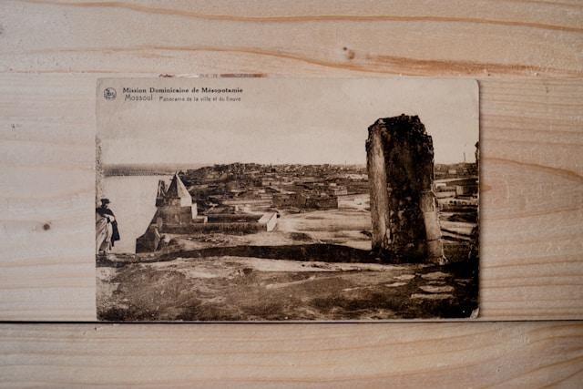 museum-of-ours-hIMg0C0yZ64-unsplash Stara sepiowa pocztówka z napisem „Mission Dominicaine de Mésopotamie – Mossoul”, przedstawiająca panoramę miasta Mosul nad rzeką, leżąca na drewnianym blacie
