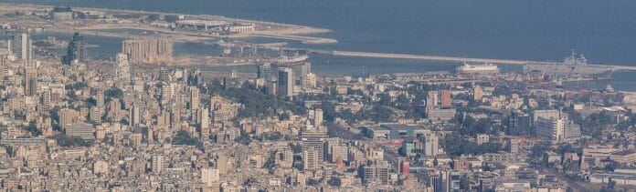 1280px-Port_of_Beirut_15-8-2020 Widok z lotu ptaka na gęsto zabudowany Bejrut i jego port nad Morzem Śródziemnym; widoczne zniszczone silosy zbożowe i gruz po wybuchu z 2020 roku.