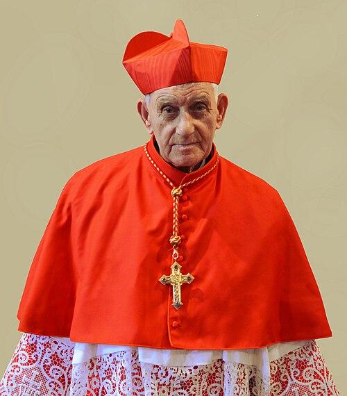 500px-Ernest_Simoni Kardynał Ernest Simoni, starszy duchowny w szkarłatnej mucecie i czerwonej piusce, ze złotym krzyżem pektoralnym na piersi, na jasnym tle