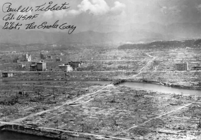 Hiroshima_aftermath Czarno-białe zdjęcie lotnicze Hiroszimy w gruzach po ataku atomowym w 1945 roku; przez zrównane z ziemią miasto wije się rzeka, w tle widać góry.