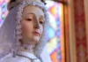 Maryja Królowa – Bóg powierzył Jej rządy nad duszami przez łaskę Zbliżenie twarzy figury Matki Bożej w białym koronkowym welonie i perłowym naszyjniku, na tle kolorowego witraża we wnętrzu kościoła.