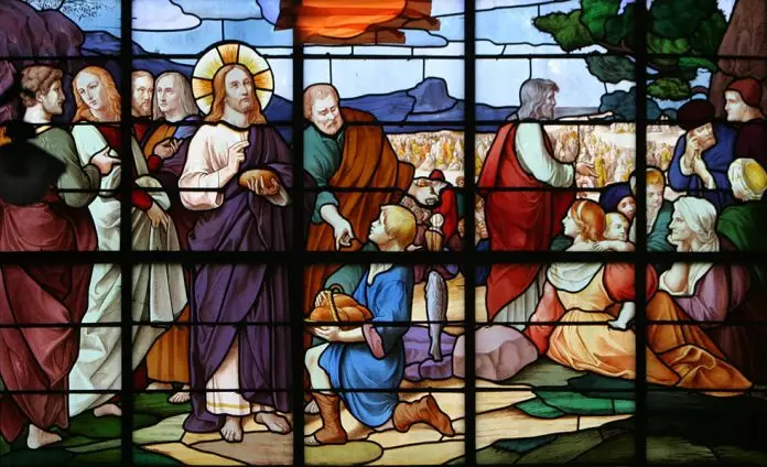 Milagre-da-multiplicacao-Parroquia-de-S-Sulpice-Fougeres-Francia-SH-mjvf-696x424-1 Witraż z cudem rozmnożenia chleba: Jezus błogosławi, klęczący chłopiec podaje bochenki chleba i ryby, obok apostołowie i siedzący tłum.