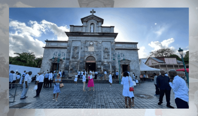 NDMCG-rrhxwhq49aq7aey90gr0gwsfrci8sqd9vwnqy4k6x4 Kamienna fasada kościoła Notre-Dame-du-Mont-Carmel w Basse-Terre na Gwadelupie z krzyżem i zegarem; przed świątynią gromadzą się odświętnie ubrani wierni