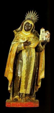 Santa_Ifigenia_-_MG_sec._XVIII Drewniana figura świętej Ifigenii — ciemnoskóra postać w czarnym płaszczu i złoconej tunice, z aureolą, trzymająca miniaturowy płonący kościół.