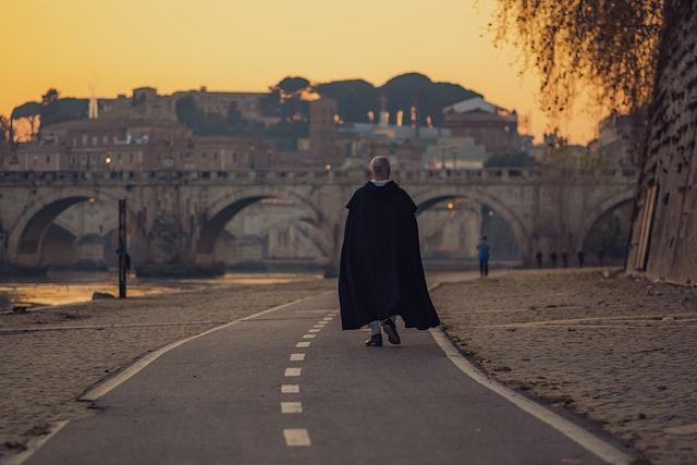 alberico-bartoccini-vl4Tb0v-pFU-unsplash Duchowny w czarnej sutannie i pelerynie idzie samotnie promenadą nad Tybrem w Rzymie o zachodzie słońca; w tle kamienny most i panorama Wiecznego Miasta.
