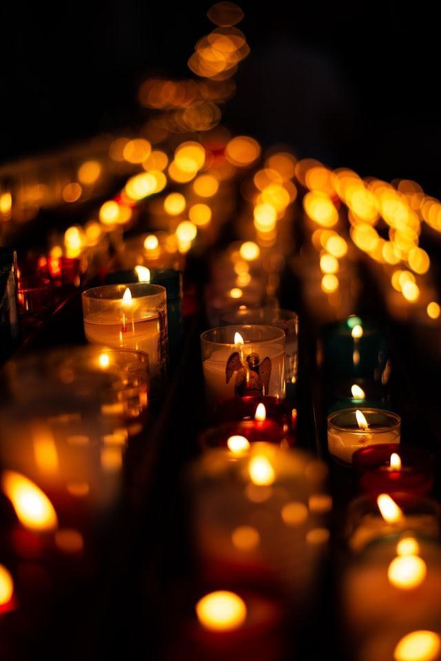aurelien-lemasson-theobald-ZwhtzQdUnSo-unsplash Rzędy zapalonych świec wotywnych jarzących się w mrocznym kościele; w jednym ze szklanych świeczników figurka anioła