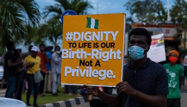 ayoola-salako-MJOtW4B2Kos-unsplash Mężczyzna w maseczce podczas protestu w Nigerii trzyma tablicę z hasłem w obronie prawa do życia oraz nigeryjską flagą