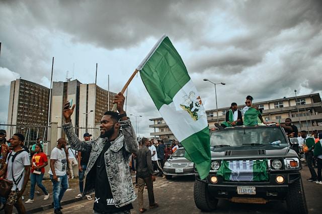emmanuel-ikwuegbu-fmPdMlgcaeY-unsplash Mężczyzna wymachujący dużą flagą Nigerii podczas ulicznego zgromadzenia; w tle udekorowany samochód terenowy i tłum na miejskiej ulicy