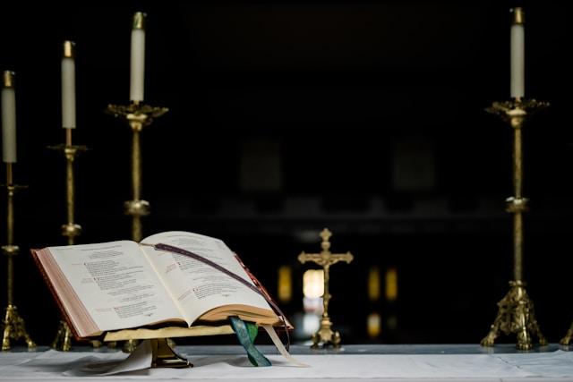 grant-whitty-nbSTmhtg7p0-unsplash-1 Otwarty mszał na złoconym pulpicie ołtarzowym, obok mosiężne świeczniki z białymi świecami, w tle krucyfiks. Zdjęcie ilustracyjne.