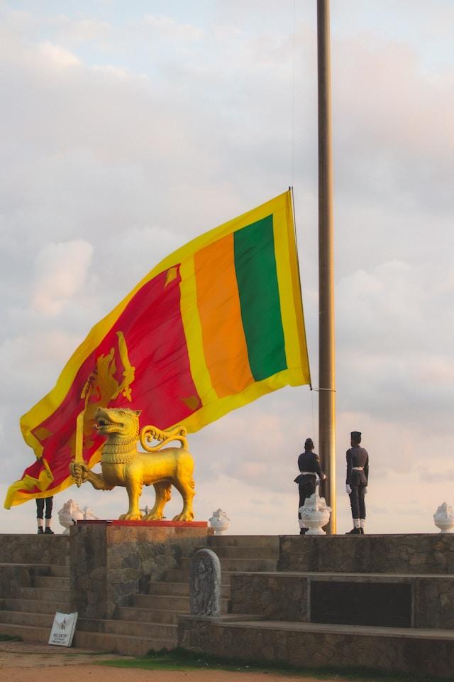 shamil-oumar-9Km4HdJCykk-unsplash Flaga narodowa Sri Lanki powiewa na maszcie nad złotym posągiem lwa, przy którym stoją dwaj umundurowani wartownicy