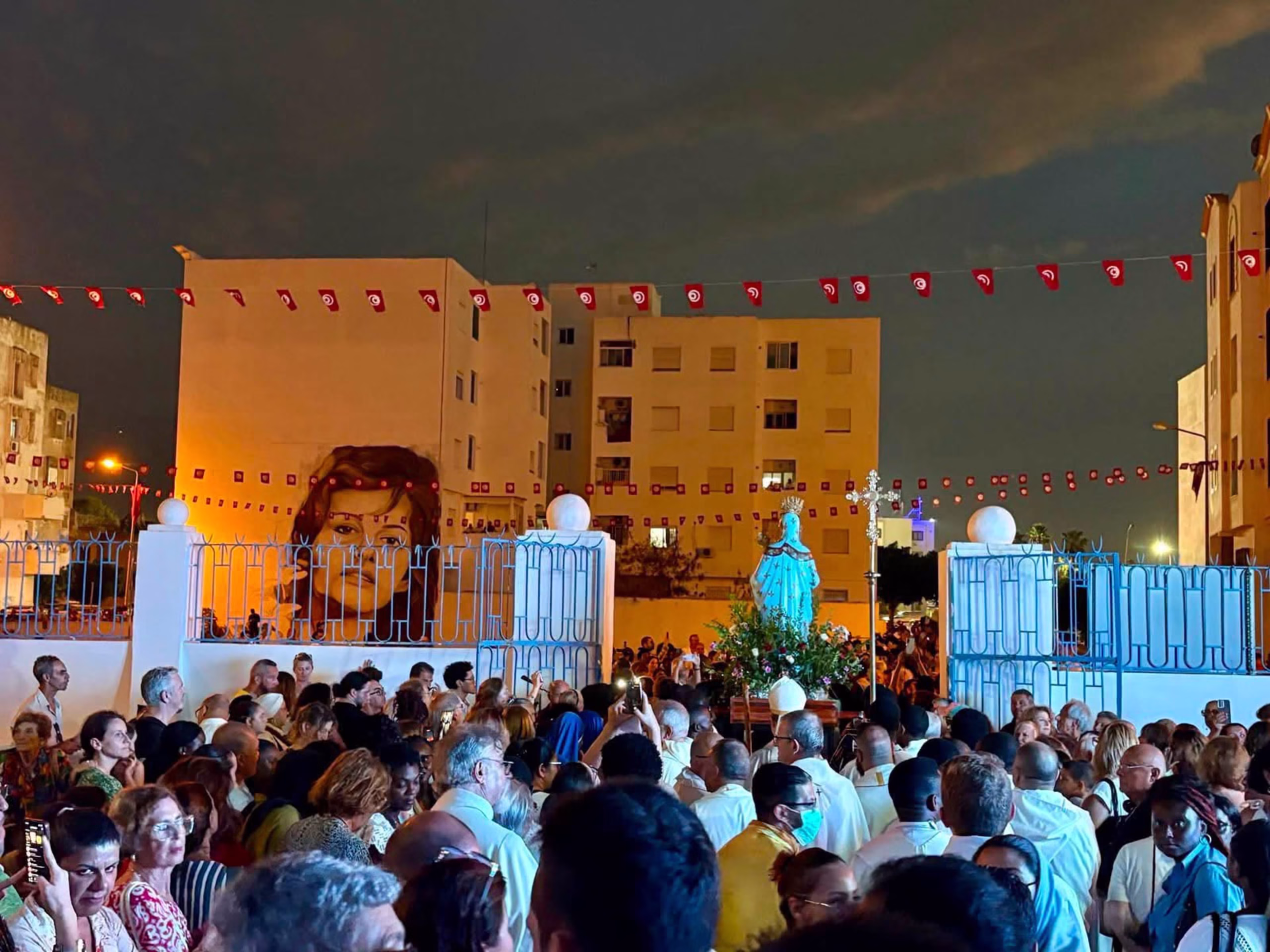 tunisia_aohlmh Wieczorna procesja Matki Bożej z Trapani w La Goulette w Tunezji: wierni niosą ukwieconą figurę Maryi, nad ulicą girlandy tunezyjskich flag.