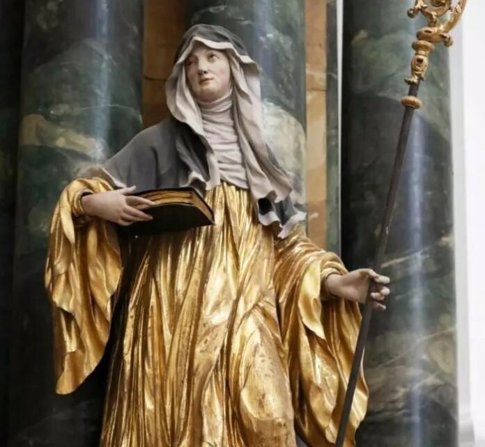 2023072616382803854_FL-700x1050-1 Złocona drewniana rzeźba św. Hildegardy z Bingen w habicie benedyktyńskim, trzymającej otwartą księgę i pastorał ksieni na tle marmurowych kolumn.