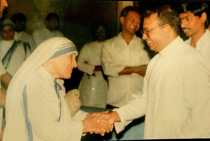20260716T0836-PRIEST-INDIA-MUSEUM-JPII-MOTHER-TERESA-1822284-1170x786-1 Matka Teresa z Kalkuty w białym sari z niebieskimi prążkami ściska dłoń uśmiechniętego księdza w okularach; w tle mężczyźni w białych koszulach