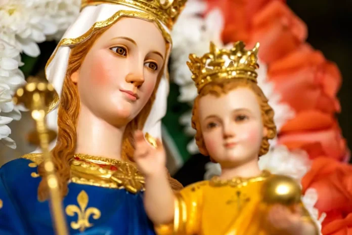 5-3-1536x1024-1 Ukoronowana figura Matki Bożej w niebieskim płaszczu w lilie, trzymająca ukoronowane Dzieciątko Jezus, otoczona białymi i czerwonymi kwiatami.