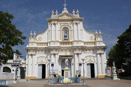 500px-Puducherry_Immaculate_Conception_Cathedral_2 Biało-złota barokowa fasada katedry Niepokalanego Poczęcia w Pondicherry, z figurą Matki Bożej w niszy i posągiem na pustym dziedzińcu; zdjęcie archiwalne.