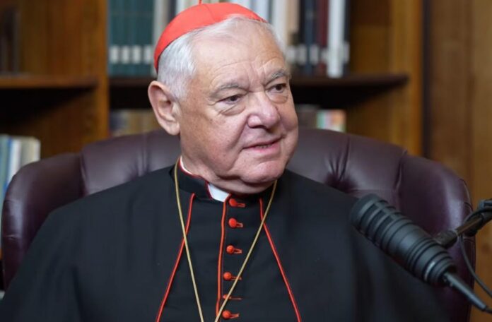 Cardinal-Muller kard. Gerhard Müller w czarnej sutannie z czerwonymi wypustkami i czerwonej piusce, siedzi w skórzanym fotelu przy mikrofonie, w tle regały z książkami.