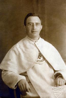 Eustaquio_van_Lieshout Sepiowy portret atelierowy bł. Eustachego van Lieshouta: kapłan w okularach, w białej pelerynie z haftowanym emblematem, siedzi w fotelu