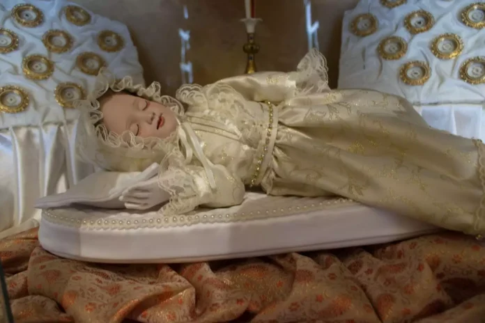 Dewocyjna figura Maryi jako śpiącego niemowlęcia, w kremowej sukni ze złotym haftem i koronkowym czepku, ułożona na białej poduszce zdobionej perłami