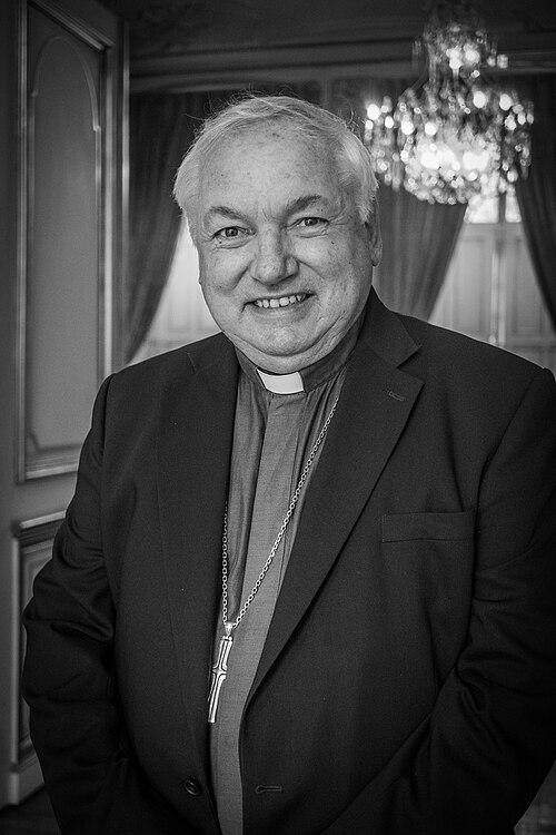 Mgr_Jean-Marc_Aveline_par_Claude_Truong-Ngoc_juillet_2021 Uśmiechnięty kard. Jean-Marc Aveline w koloratce i ciemnej marynarce, z krzyżem pektoralnym – czarno-biały portret we wnętrzu z żyrandolem.