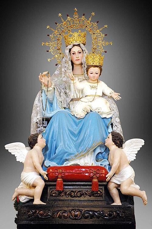 Our_Lady_of_Arabia_-_Pontifically_crowned_in_Kuwait Polichromowana figura Matki Bożej Arabskiej z Dzieciątkiem Jezus na tronie, oboje w złotych koronach, Maryja z różańcem, u stóp dwaj aniołkowie.