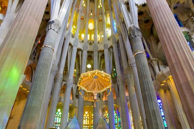 aki-A27sA6kViXc-unsplash Wnętrze bazyliki Sagrada Família w Barcelonie, z kolumnami przypominającymi drzewa i baldachimem nad ołtarzem.