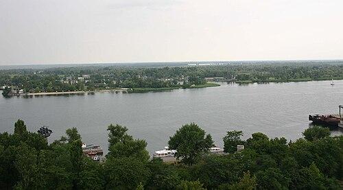 500px-Ukraine_Dnepr_bei_Cherson Szeroka rzeka Dniepr widziana z góry, z zalesionymi brzegami i zabudowaniami w oddali; przy brzegu łodzie i barka