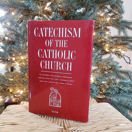 Catechism_of_the_Catholic_Church_in_front_of_a_tree Czerwony tom „Catechism of the Catholic Church” (wydanie CCCB) na wiklinowym siedzisku na tle choinki z białymi lampkami