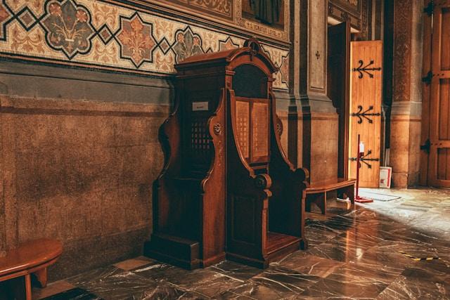 Konfesjonał Rzeźbiony drewniany konfesjonał z kratkami przy ścianie kościoła zdobionej ornamentem, obok otwarte drewniane drzwi i lśniąca marmurowa posadzka