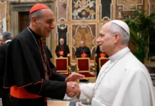Po „Venite ad me”: czy Leon XIV na nowo zdefiniuje watykański urząd doktrynalny? Papież Leon XIV w białej sutannie ściska dłoń kard. Víctora Manuela Fernándeza w bogato zdobionej sali; w tle siedzą duchowni w sutannach.