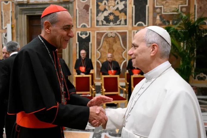 Fernandez-and-Leo Papież Leon XIV w białej sutannie ściska dłoń kard. Víctora Manuela Fernándeza w bogato zdobionej sali; w tle siedzą duchowni w sutannach.