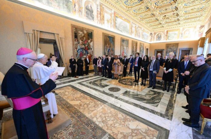 Papież Leon XIV z uniesioną ręką błogosławi uczestników spotkania stojących w zdobionej freskami sali w Watykanie; obok duchowny w fioletowej piusce