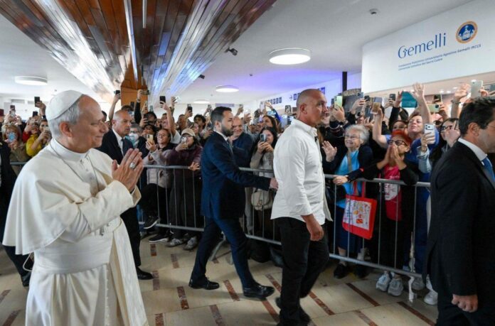 Papież Leon XIV w białej sutannie składa dłonie, idąc holem Polikliniki Gemelli obok tłumu za barierkami, który fotografuje go telefonami