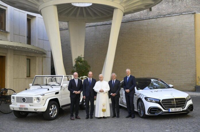 Papież Leon XIV z czterema mężczyznami w garniturach między białym, otwartym papamobile Mercedes-Benz z tablicą SCV 1 a białą limuzyną Mercedes-Benz