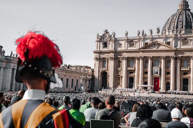 michael-hunter-HKEI512Qefo-unsplash Gwardzista szwajcarski w hełmie z czerwonym pióropuszem, w tle tłum wiernych na placu św. Piotra i fasada bazyliki watykańskiej